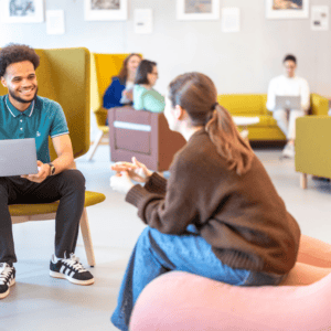 Espace commun détente coworking : personnes en conversation, canapés, ordinateurs portables, ambiance étudiante.