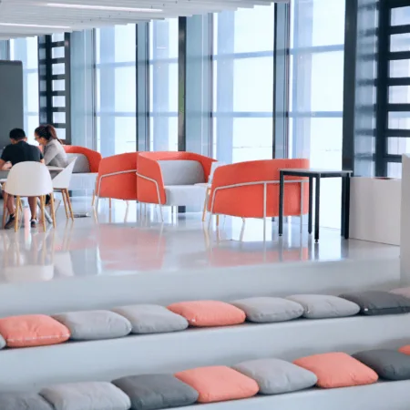 Espace communal moderne avec banquettes, coussins corail, fauteuils et larges baies vitrées.