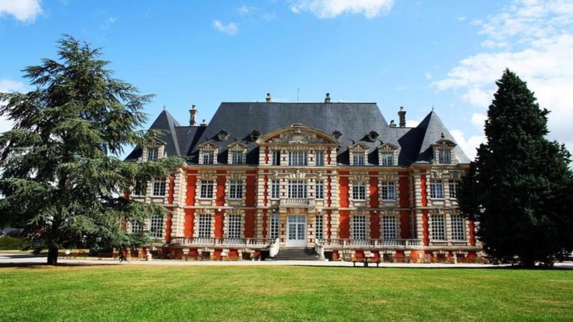 Façade de manoir en briques et pierre, toit dardoise, pelouse et arbres.