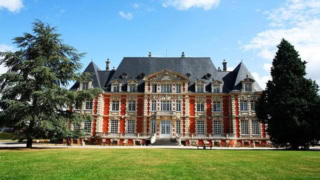 Façade de manoir en briques et pierre, toit dardoise, pelouse et arbres.