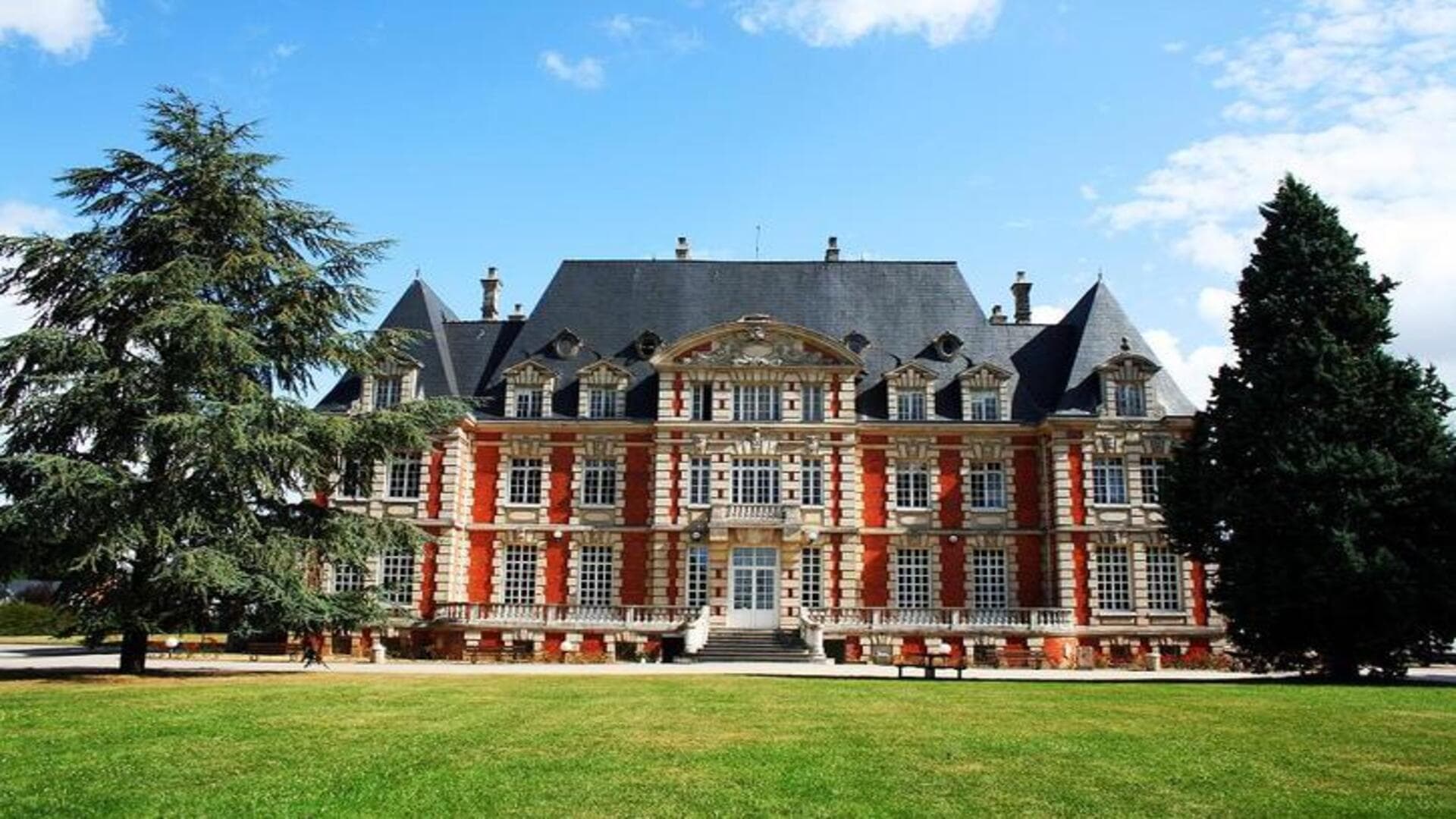 Façade de manoir en briques et pierre, toit dardoise, pelouse et arbres.