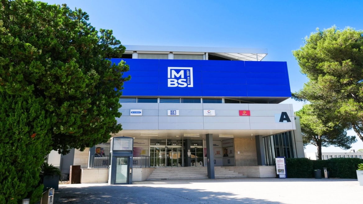 Façade moderne avec entrée, grand panneau bleu avec logo et escalier, bâtiment public entouré darbres.