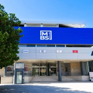 Façade moderne avec entrée, grand panneau bleu avec logo et escalier, bâtiment public entouré darbres.