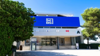 Façade moderne avec entrée, grand panneau bleu avec logo et escalier, bâtiment public entouré darbres.