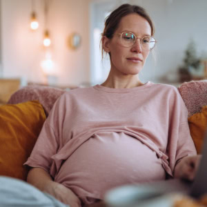 Femme enceinte sur canapé avec ordinateur portable, lunettes, ambiance cosy et lumière naturelle.