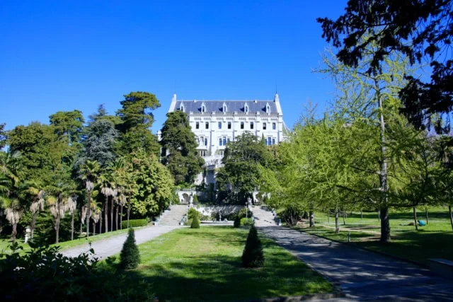 Manoir blanc historique entouré de jardins, fontaine, palmiers et pelouses sous ciel bleu.