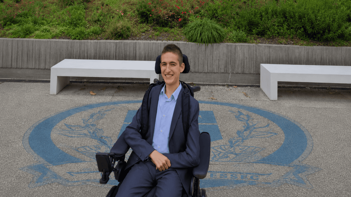 Jeune homme en fauteuil roulant électrique en costume, portrait souriant sur campus extérieur.
