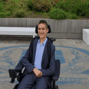 Jeune homme en fauteuil roulant électrique en costume, portrait souriant sur campus extérieur.