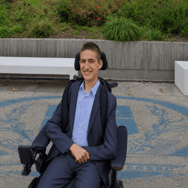 Jeune homme en fauteuil roulant électrique en costume, portrait souriant sur campus extérieur.