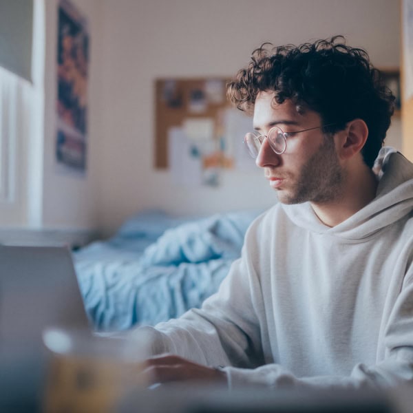Jeune homme étudiant avec ordinateur portable dans chambre lumineuse, lunettes et concentration.