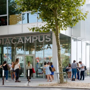 Entrée MediaCampus : jeunes étudiants devant bâtiment moderne à façade vitrée, campus urbain.
