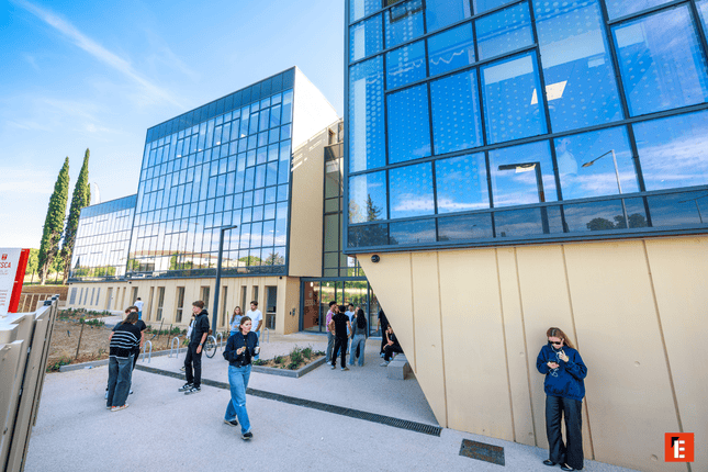 Cour extérieure moderne entre bâtiments en verre, façades réfléchissantes, personnes, architecture contemporaine ensoleillée.