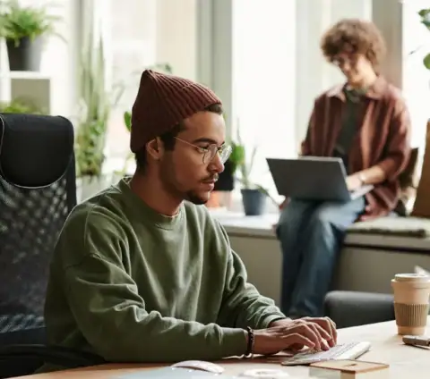Personne travaillant au bureau en coworking, ordinateur, café, plantes et lumière naturelle.