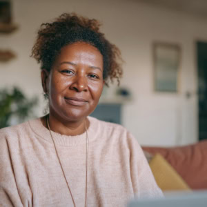 Portrait femme souriante en pull beige, intérieur cosy, lumière naturelle.