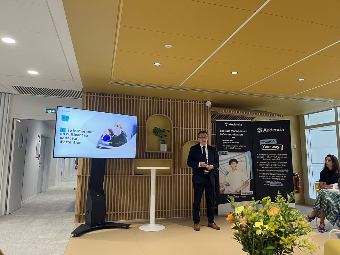 Audencia : conférencier en costume dans salle de présentation moderne avec écran et décor bois.