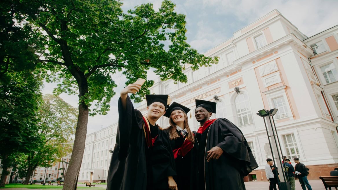 Remise de diplômes : trois étudiants en toges prennent un selfie devant un bâtiment universitaire.