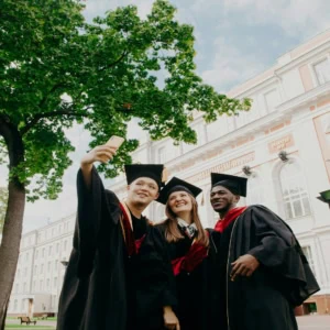 Remise de diplômes : trois étudiants en toges prennent un selfie devant un bâtiment universitaire.