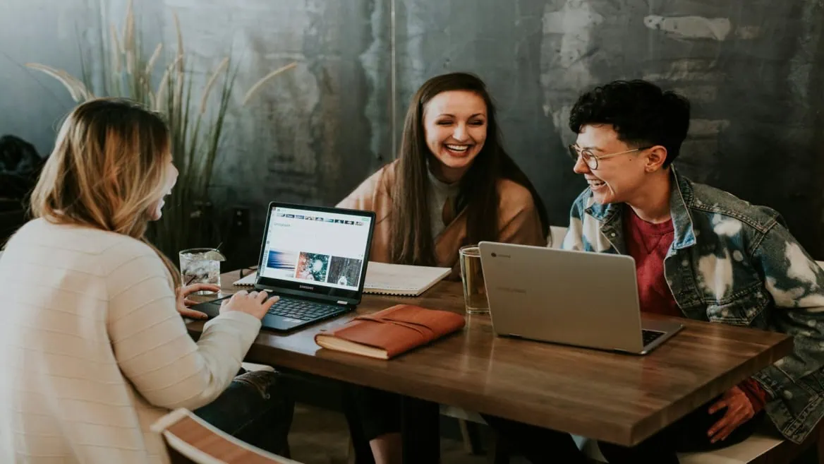 Réunion créative de trois personnes riant autour dordinateurs dans un espace coworking chaleureux.