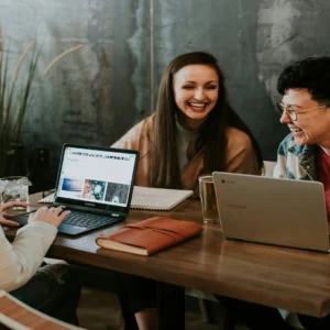 Réunion créative de trois personnes riant autour dordinateurs dans un espace coworking chaleureux.