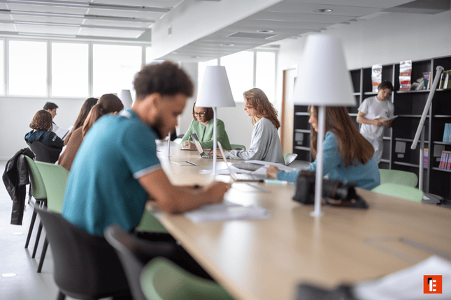 Salle détude lumineuse avec longue table, étudiants, lampes de bureau et étagères de livres.