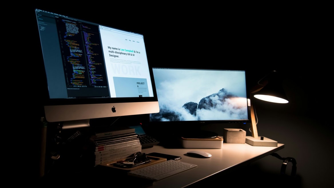 Bureau informatique nocturne pour développeur designer avec iMac, double écran, clavier, lampe, code et portfolio.