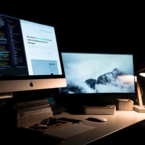 Bureau informatique nocturne pour développeur designer avec iMac, double écran, clavier, lampe, code et portfolio.