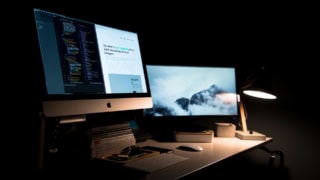Bureau informatique nocturne pour développeur designer avec iMac, double écran, clavier, lampe, code et portfolio.