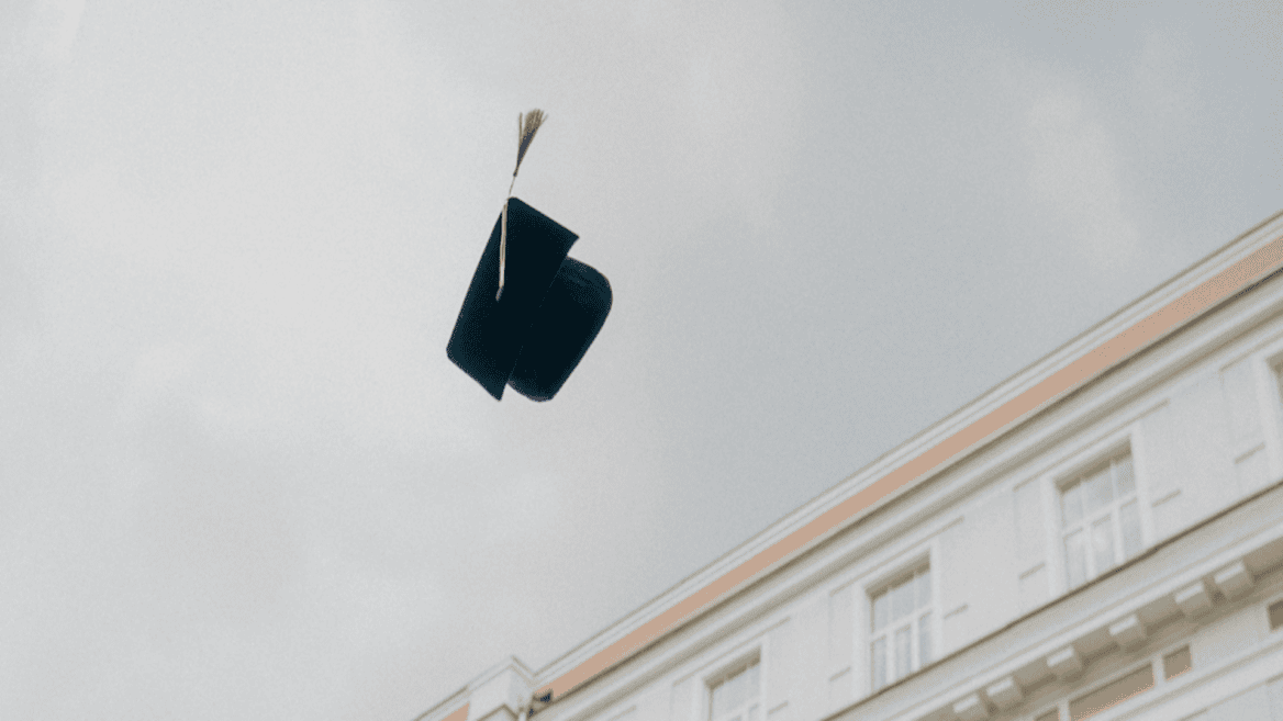 Chapeau de diplômé noir avec gland doré lancé dans le ciel au-dessus dun bâtiment classique.