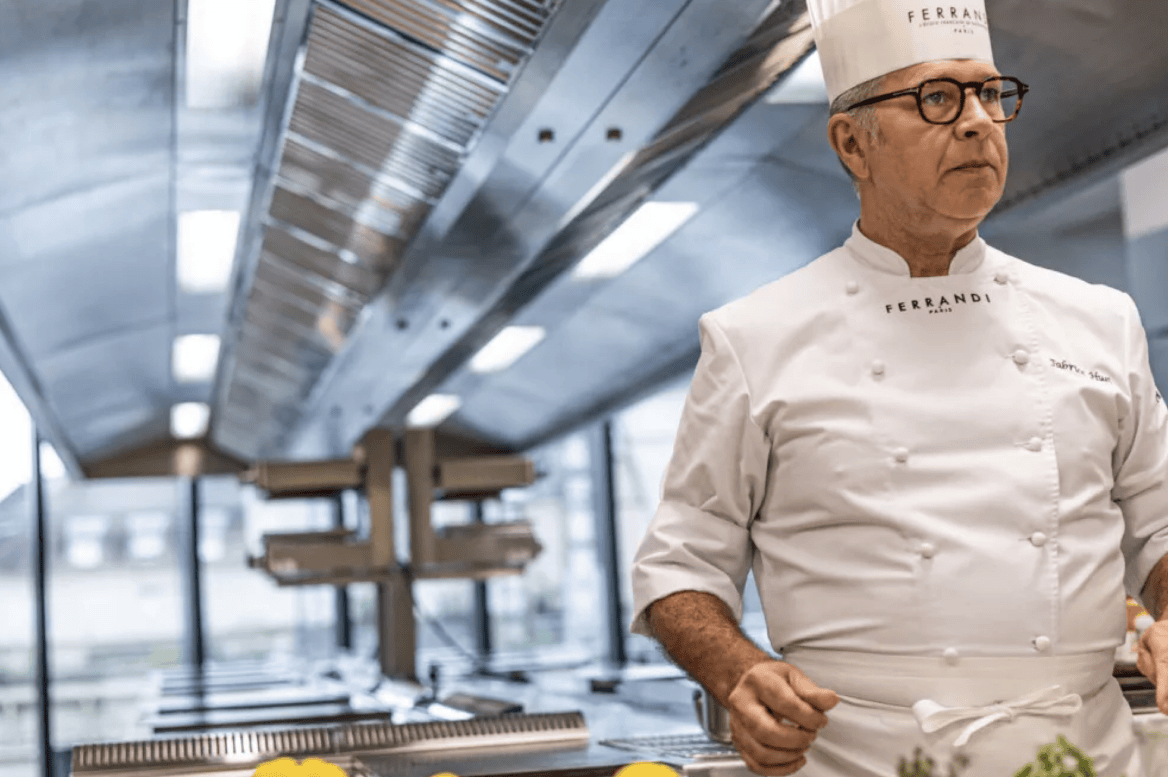 Chef en toque et veste blanche dans cuisine professionnelle industrielle, citrons sur plan de travail.