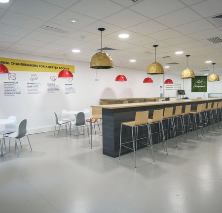 Cuisine commune moderne avec grande table centrale, tabourets, kitchenette et lampes design.