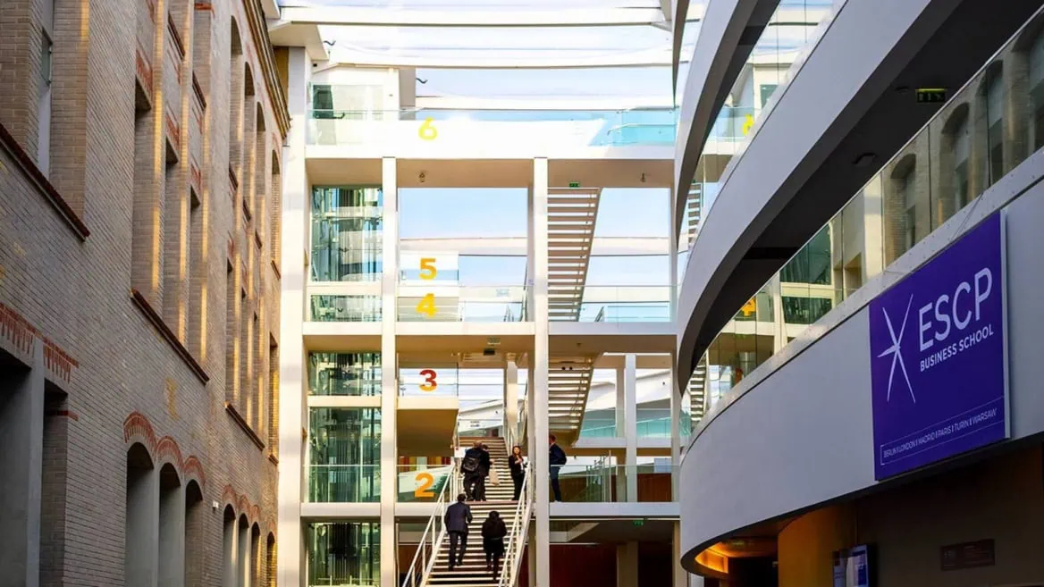 Atrium lumineux ESCP Business School avec escalier central, verrière, mur en brique et façade vitrée.