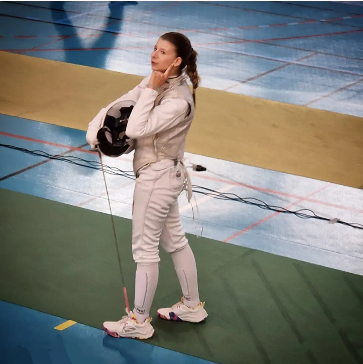 Escrimeuse en tenue blanche tenant masque et épée sur piste de compétition.