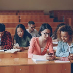 Étudiants en amphithéâtre universitaire partageant un smartphone et prenant des notes.
