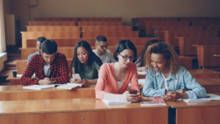 Étudiants en amphithéâtre universitaire partageant un smartphone et prenant des notes.