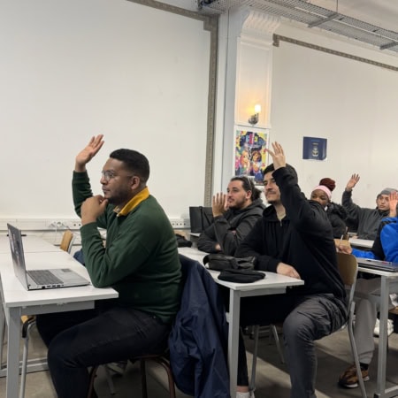 Étudiants en classe levant la main, ordinateurs portables et ambiance studieuse.
