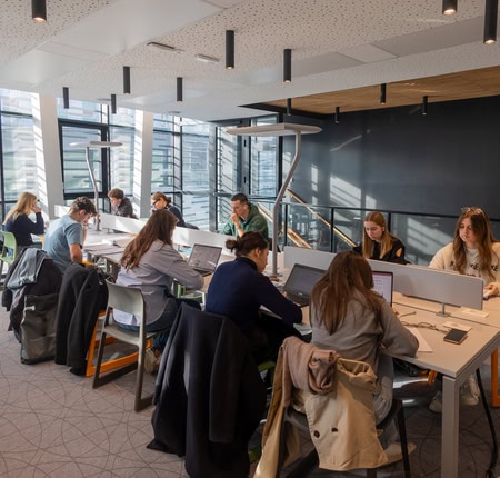 Espace de coworking lumineux avec grandes fenêtres, postes de travail partagés, personnes travaillant.