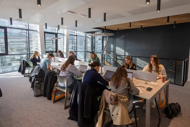 Espace de coworking lumineux avec grandes fenêtres, postes de travail partagés, personnes travaillant.
