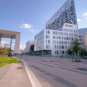 Place urbaine moderne avec arche monumentale, bâtiments contemporains, espaces piétonniers et art urbain.