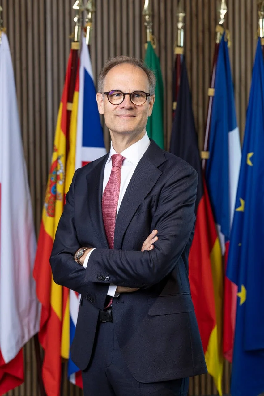 Portrait professionnel dun homme en costume, cravate et lunettes devant des drapeaux nationaux.