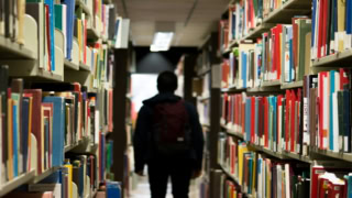 Allée de bibliothèque bordée de rayonnages remplis de livres, silhouette détudiant au loin.