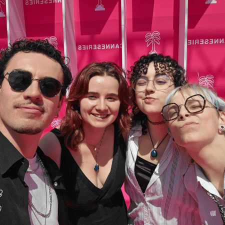 Selfie quatre amis devant fond rose ensoleillé, sourires, lunettes et bijoux.