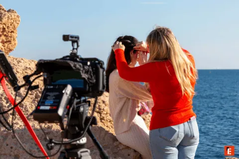 Tournage plage : caméra sur rochers, équipe coiffure préparant actrice devant mer.