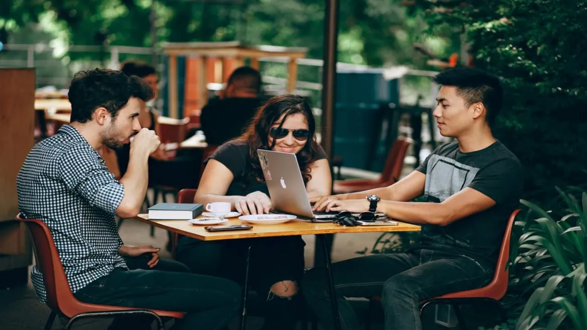 Trois personnes en terrasse de café autour d’un ordinateur portable, conversation et collaboration.