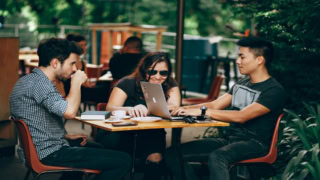 Trois personnes en terrasse de café autour d’un ordinateur portable, conversation et collaboration.