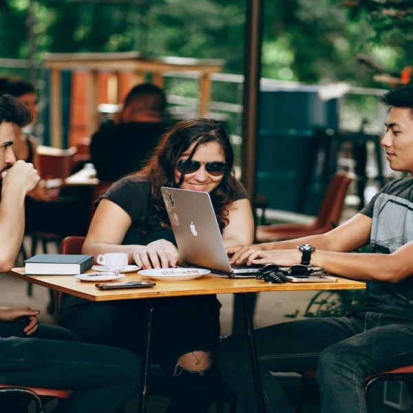 Trois personnes en terrasse de café autour d’un ordinateur portable, conversation et collaboration.