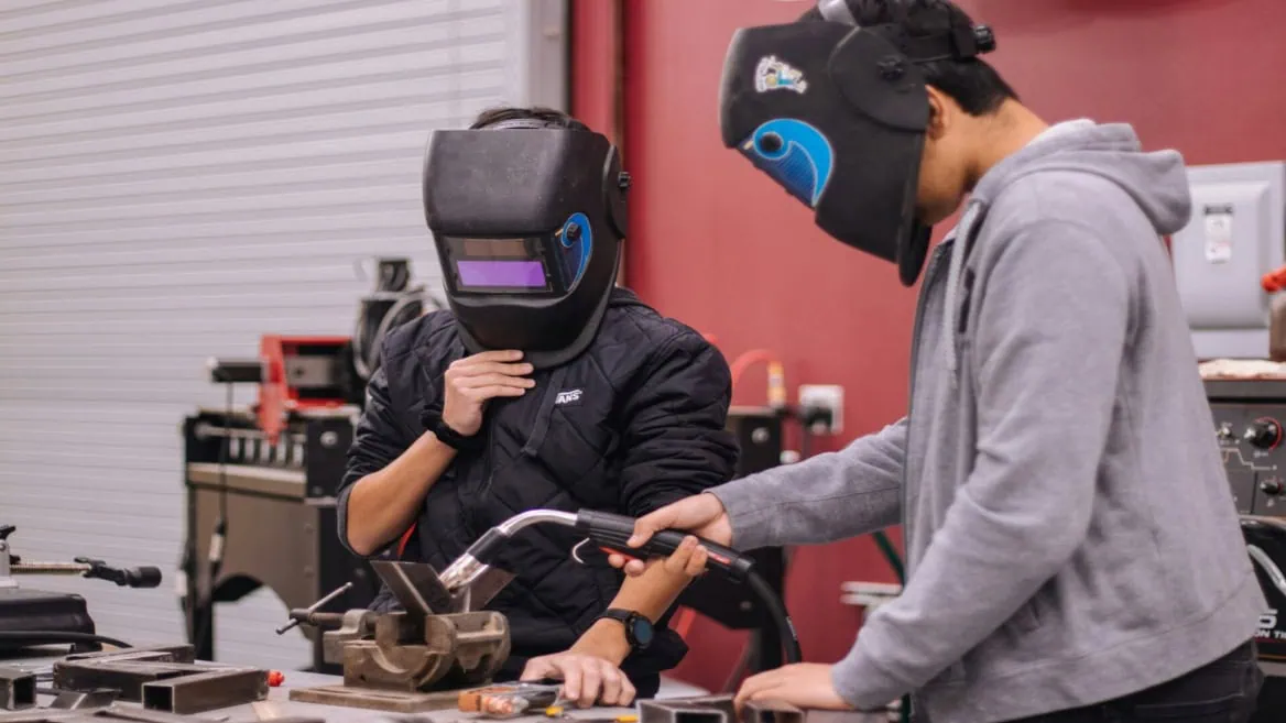 Deux personnes en atelier de soudure avec casques, étau et torche sur établi.