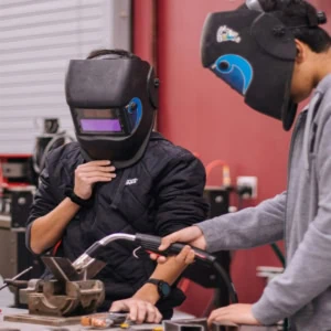 Deux personnes en atelier de soudure avec casques, étau et torche sur établi.