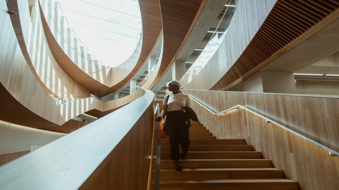 Atrium moderne avec escalier en bois, balustrades en verre et personne descendant sous lumière naturelle.