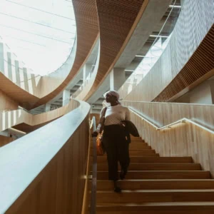 Atrium moderne avec escalier en bois, balustrades en verre et personne descendant sous lumière naturelle.