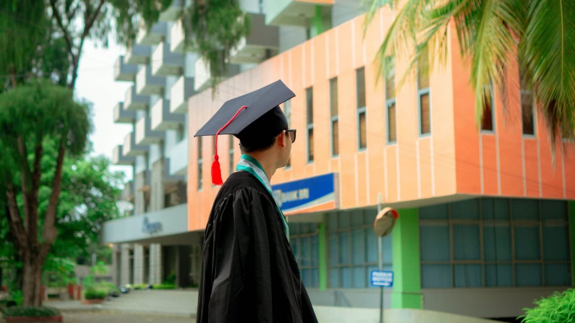 Étudiant diplômé vu de dos en toge noire, toque à pompon rouge, sur campus urbain.